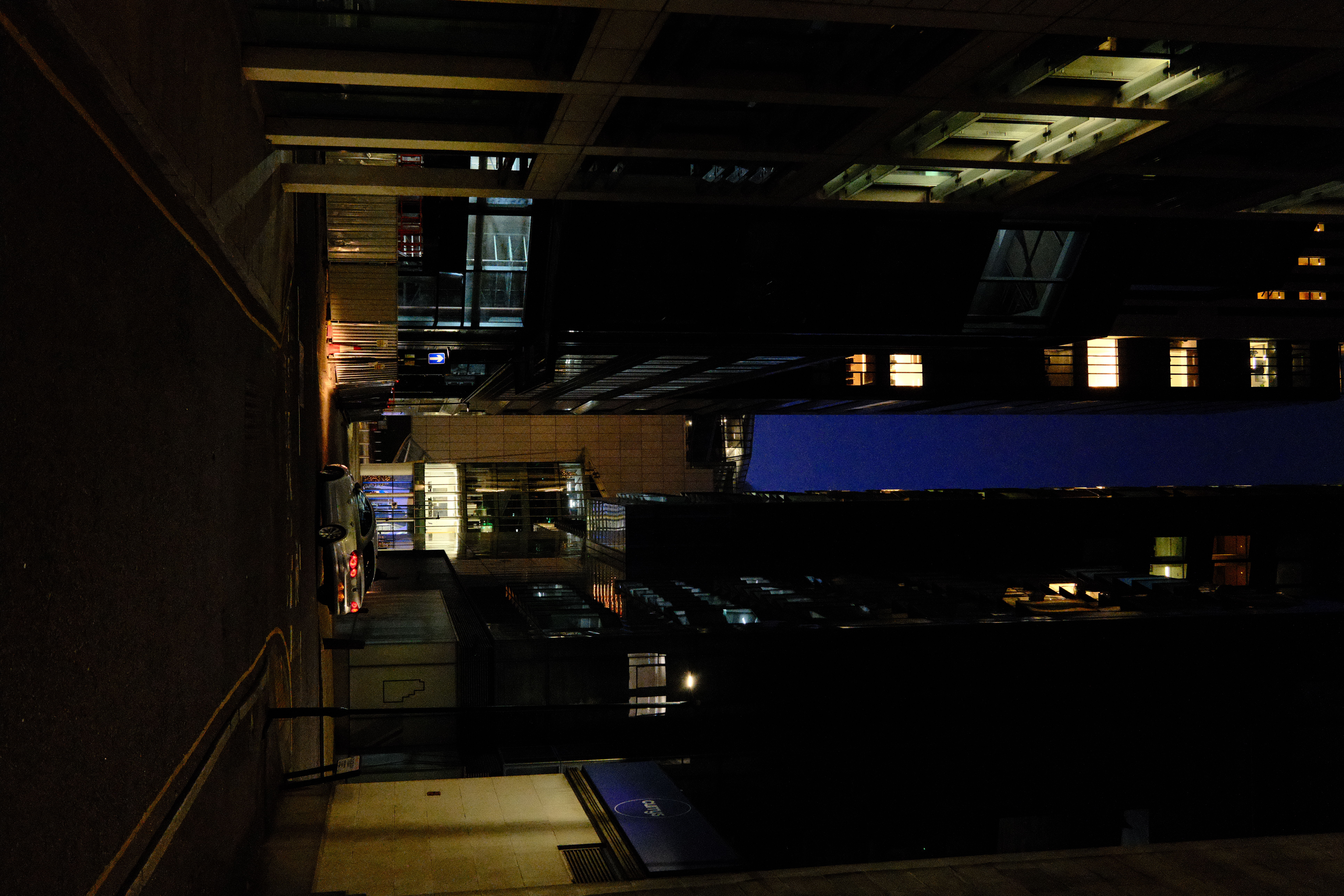 A narrow city alleyway at night, tall office buildings either side, a sliver of blue sky above
