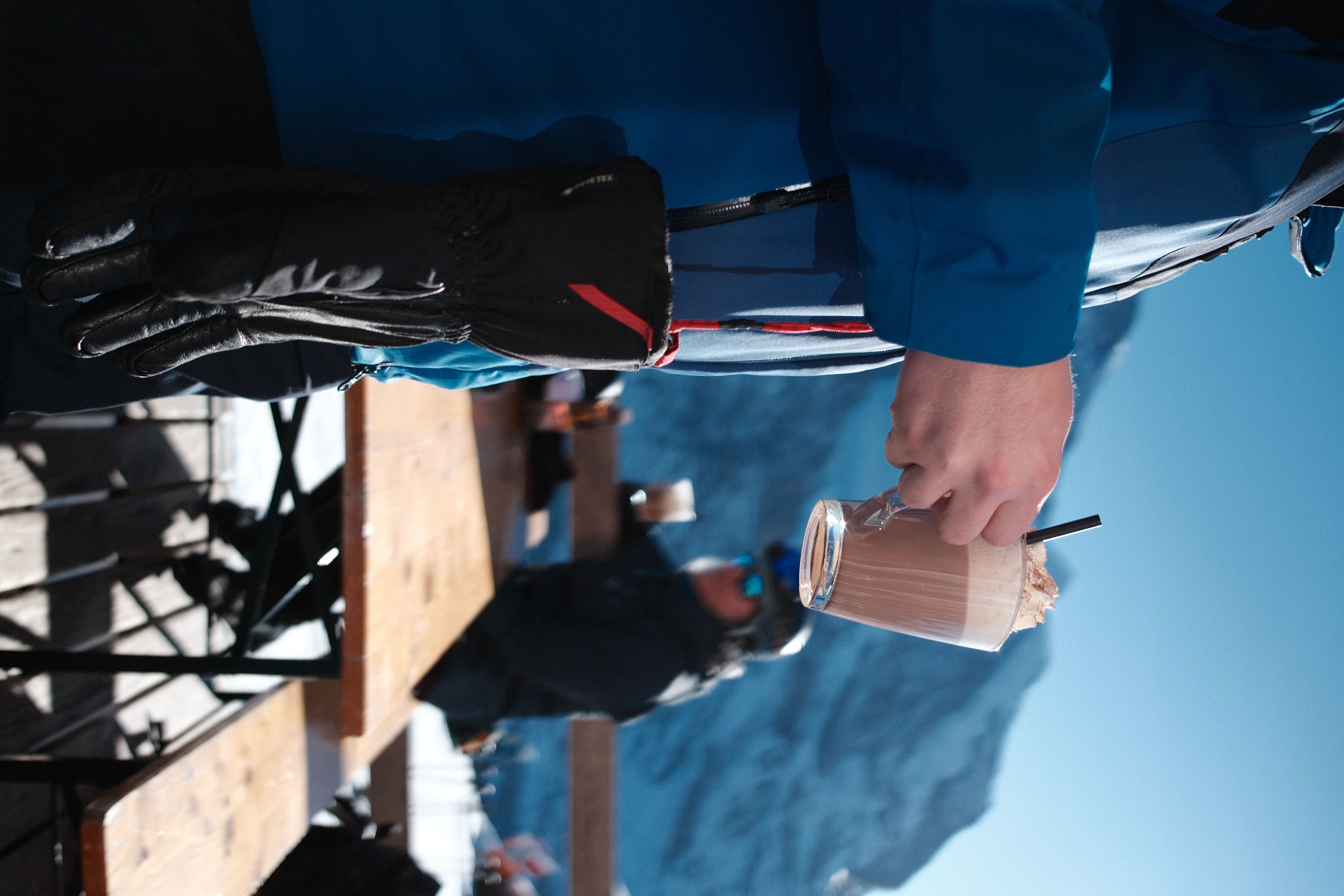 A person in ski gear holding a hot chocolate on a mountain terrace, snowy peaks behind