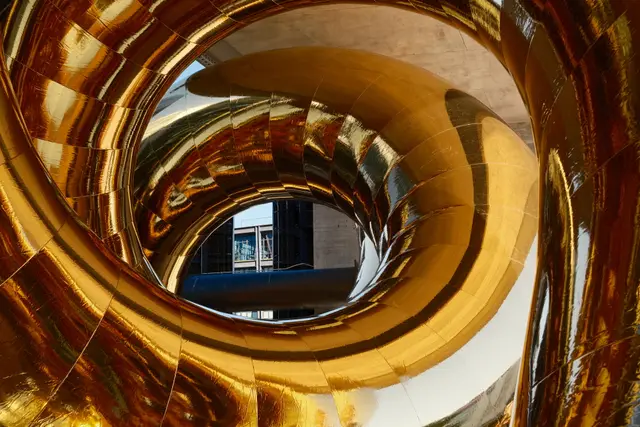 A towering polished gold tubular sculpture reflecting the plaza and passers-by
