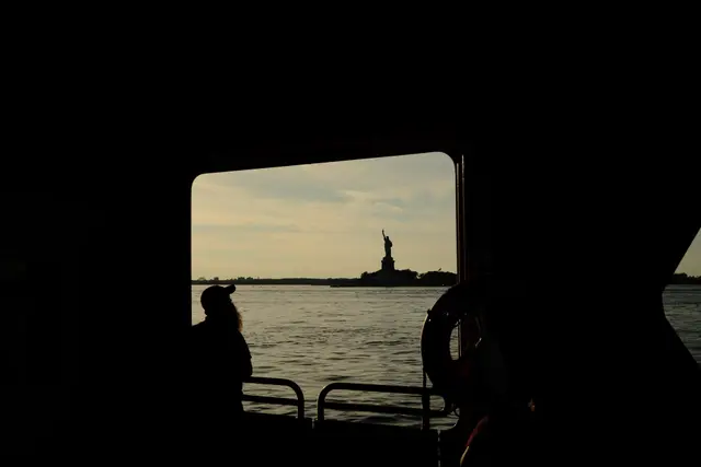 The Statue of Liberty silhouetted through a ferry window, a passenger and lifebuoy in the foreground